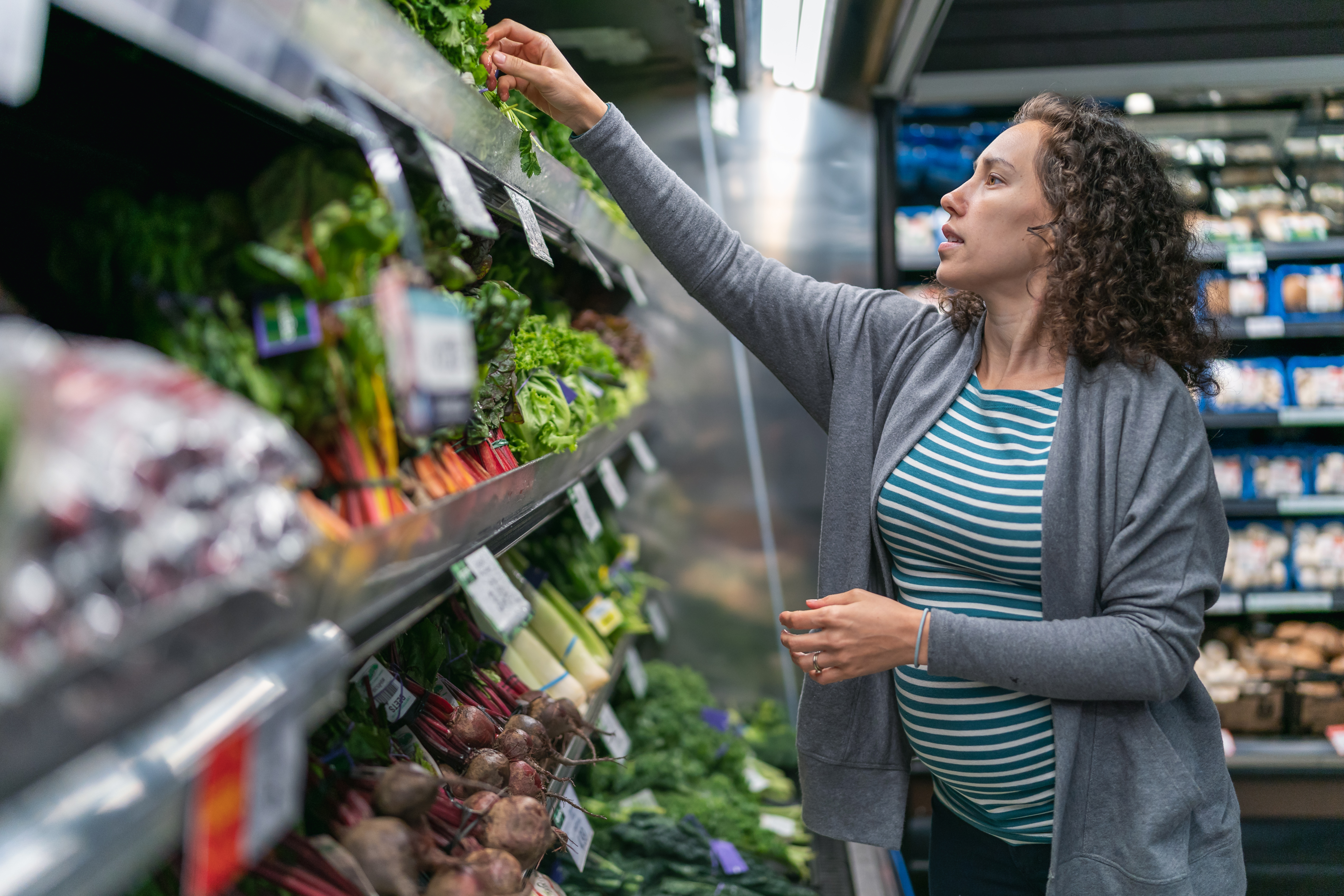 A young mixed race pregnant woman is at the grocery store. The healthy woman is buying fresh fruit and vegetables. She is reaching for fresh herbs in the refrigerated area of the store's produce section. She is wearing casual maternity clothing.